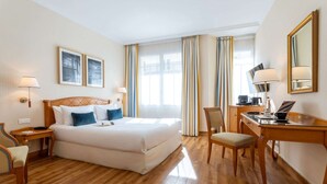 Habitación de hotel con cama, escritorio, silla y un amplio ventanal con cortinas.