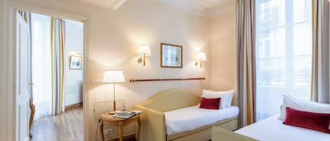 A hotel room with a bed, a bedside table, a lamp, a window with curtains, and a door leading to another room.