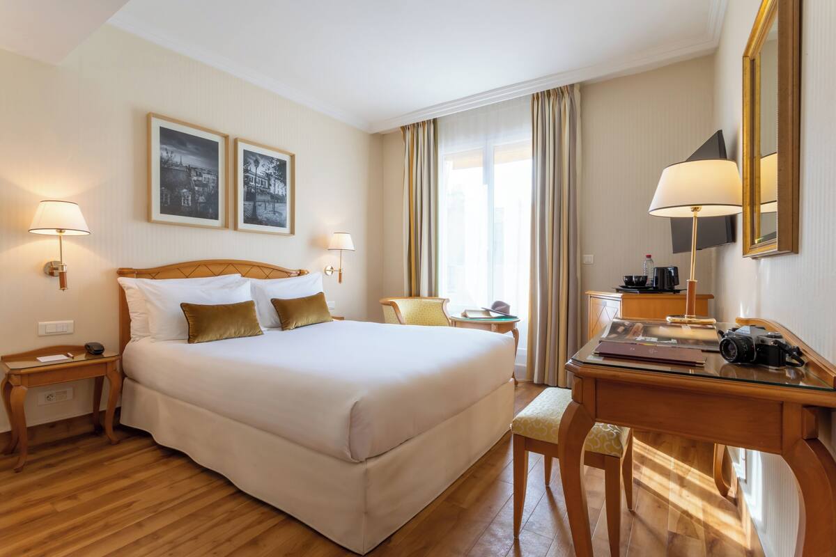 A hotel room with a large bed, two bedside lamps, a desk with a camera, a chair, and a small table.