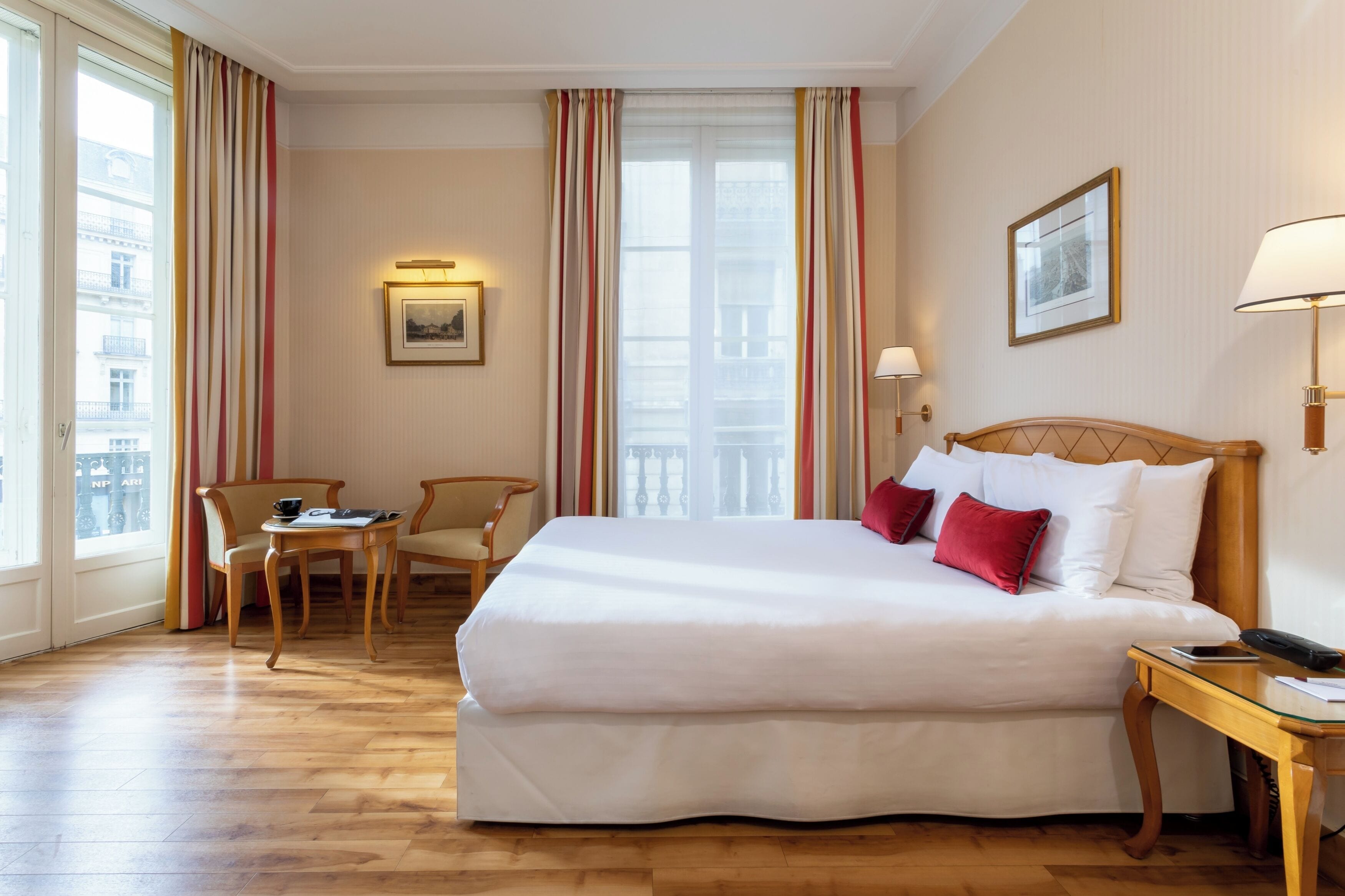 A hotel room with a large bed, a small table, two chairs, a nightstand, and a window with curtains.