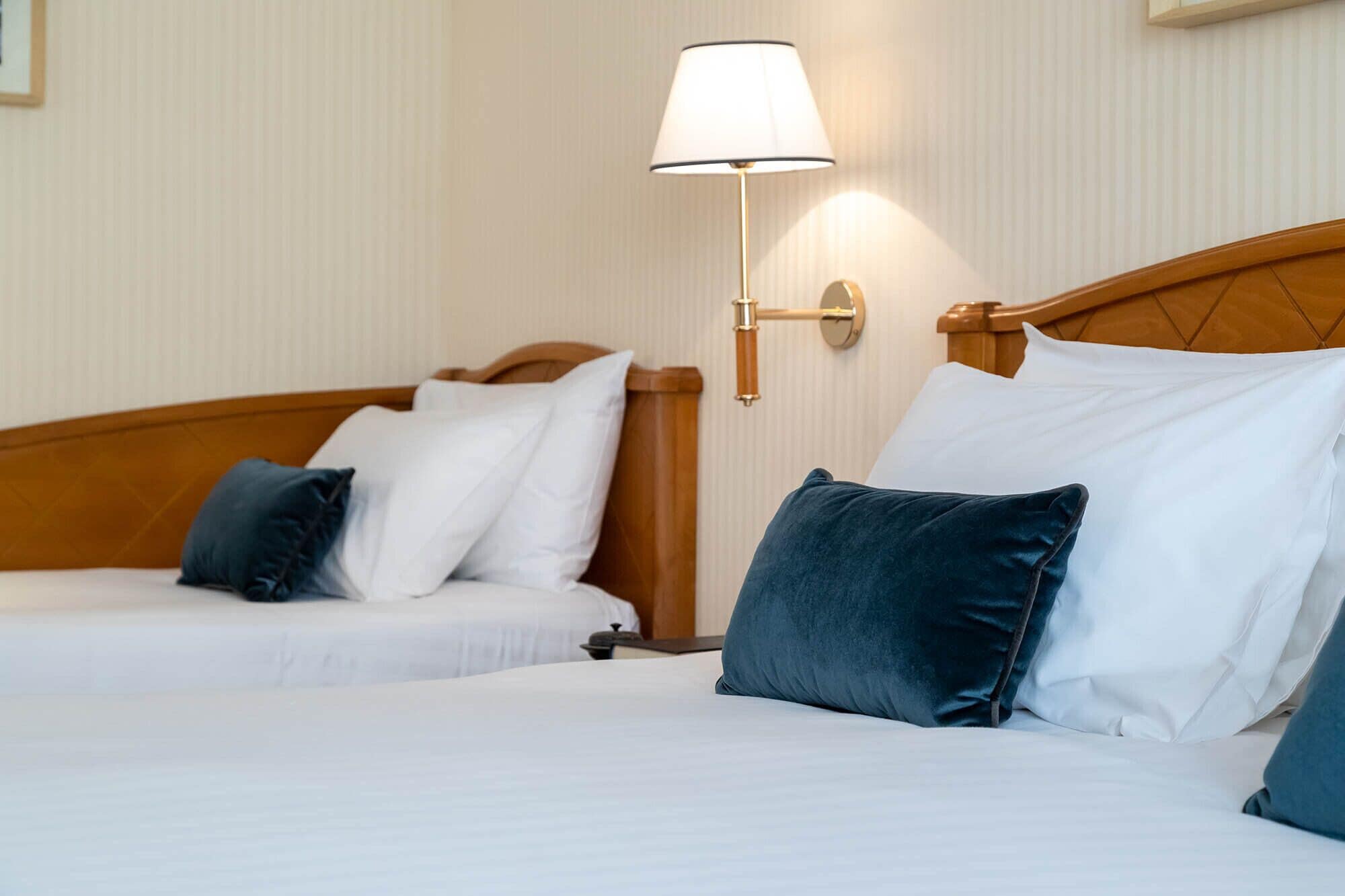 A hotel room with two beds, each with white bedding and blue pillows. There is a wall-mounted lamp with a white shade.