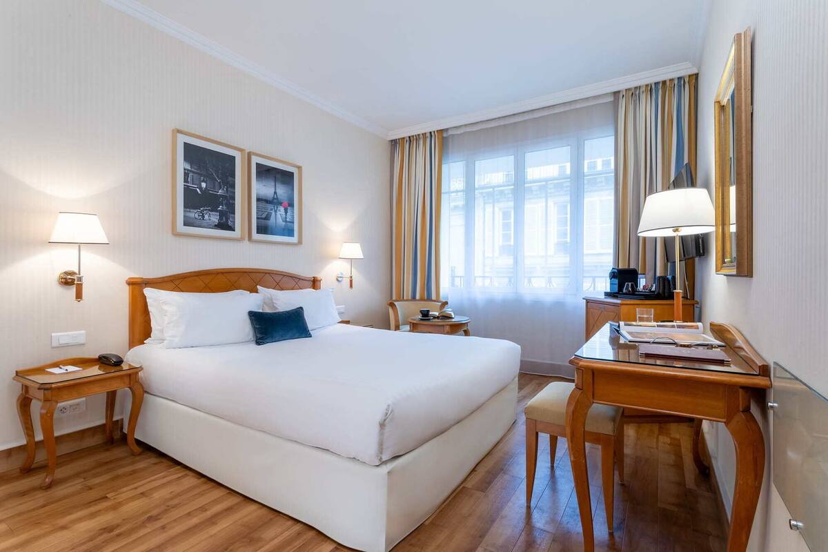 A hotel room with a large bed, two bedside tables, a desk, and a chair. There are two lamps, a window with curtains, and framed pictures on the wall.