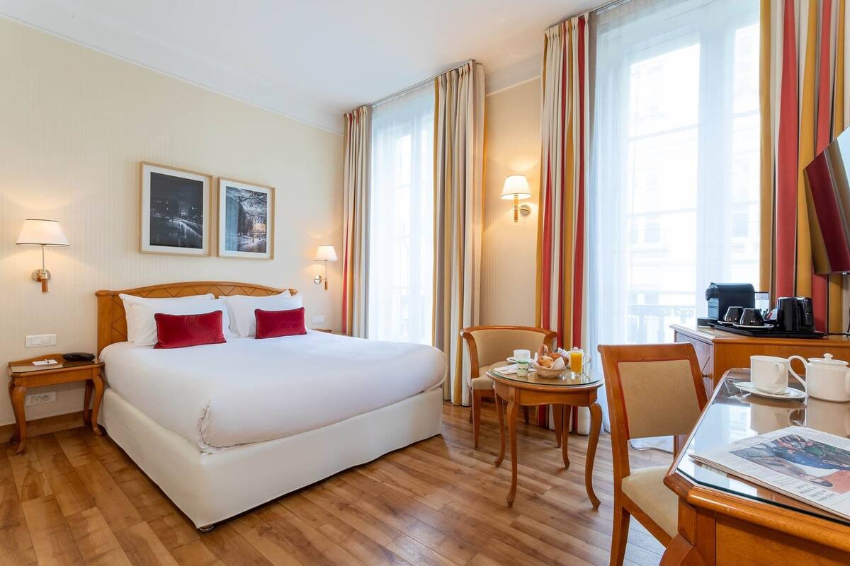 A hotel room with a large bed, a desk with a chair, a small table with a chair, and a window with curtains.