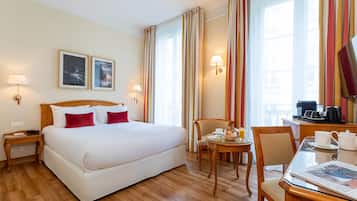 Una habitación de hotel con una cama grande, un escritorio con silla, una mesita con otra silla y una ventana con cortinas.