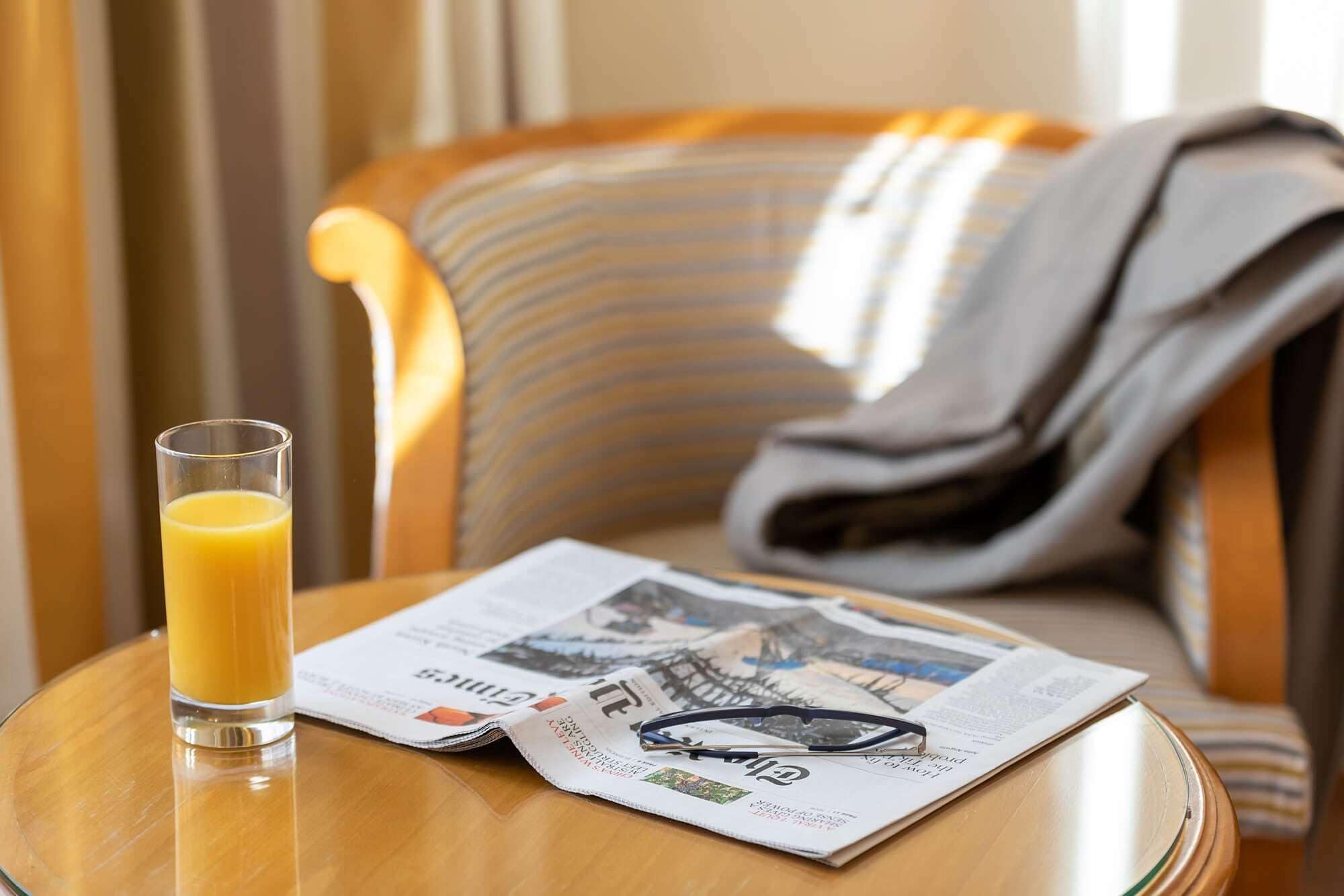 A glass of orange juice on a round table with a newspaper and glasses.
