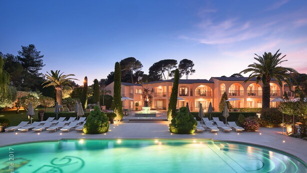 A large, well-lit swimming pool with lounge chairs and umbrellas, surrounded by manicured landscaping and a grand building with multiple balconies. - Villa Miraé by Inwood Hotels - Cap d'Antibes (Antibes)