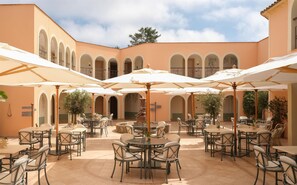 An outdoor cafe with multiple tables and chairs, surrounded by arches and covered by umbrellas.