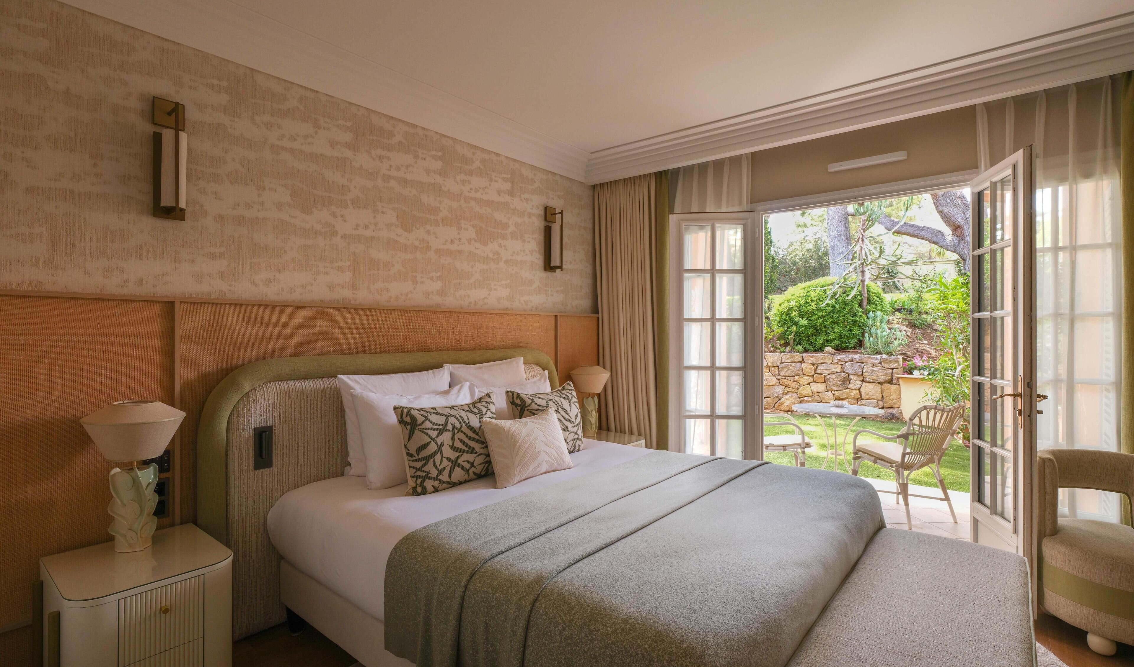 Ein Hotelzimmer mit einem großen Bett, einem Nachttisch, einer Lampe und Blick auf einen Garten durch eine Schiebetür.