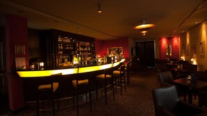 A dimly lit bar area with a bar counter, stools, and a well-stocked shelf with bottles. - Turm Hotel (Frankfurt)