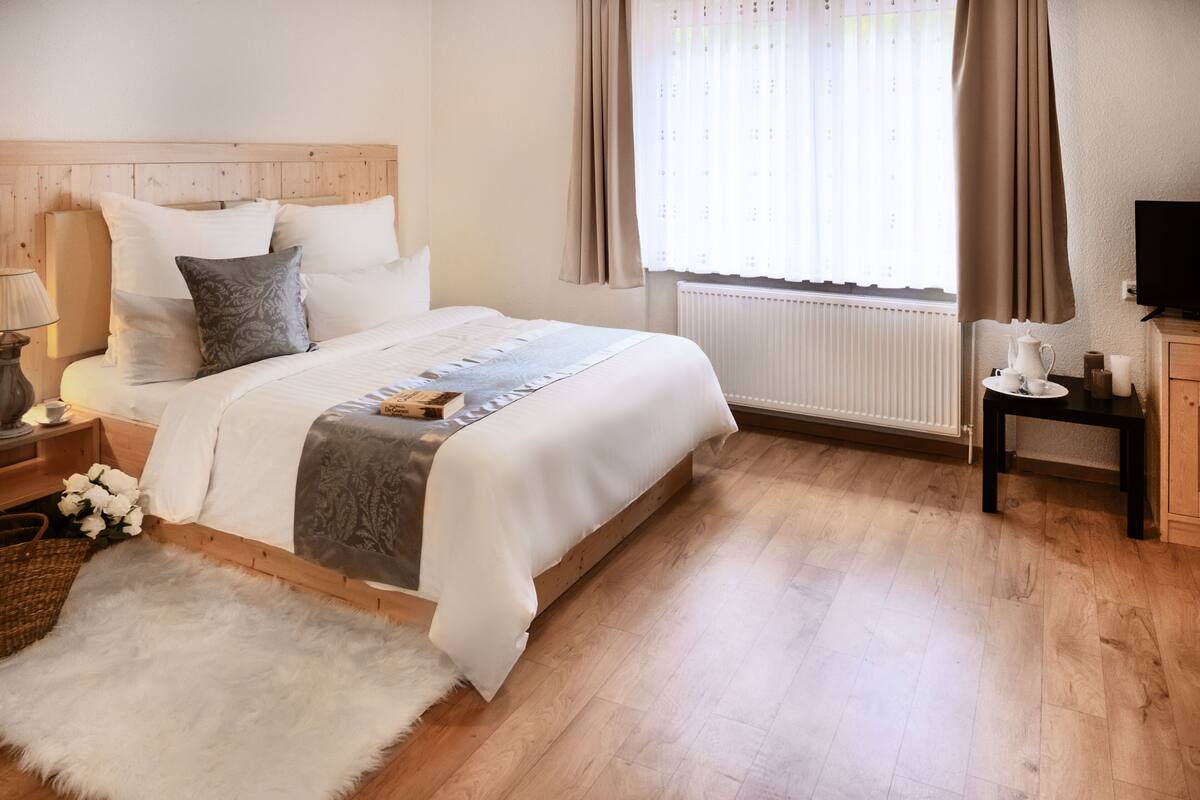 Ein ordentlich eingerichtetes Schlafzimmer mit einem Holzkopfteil, einer weißen Bettdecke, einem flauschigen weißen Teppich und einem Nachttisch mit einer Lampe.