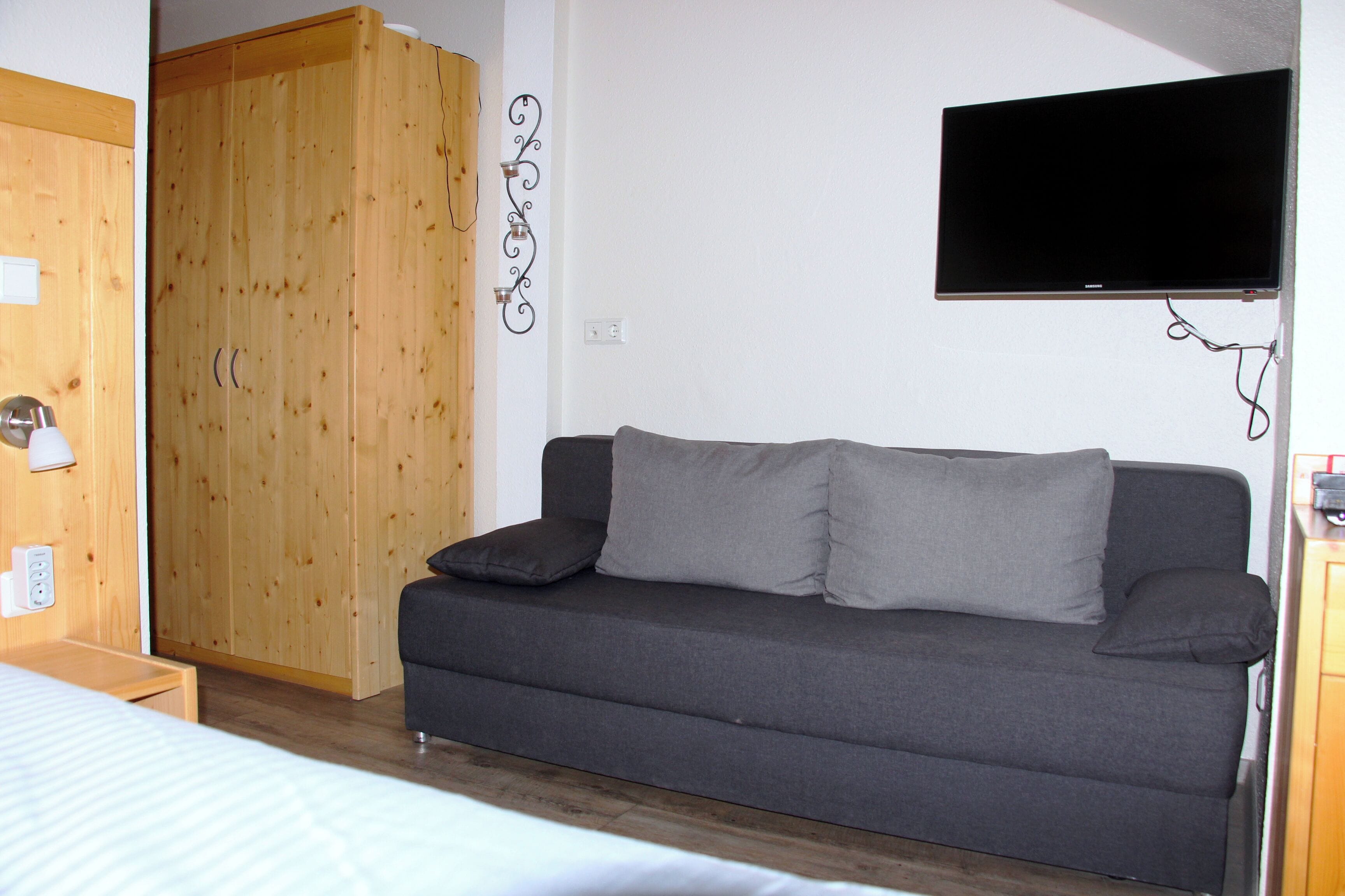 A room with a grey sofa, a wooden wardrobe, a television mounted on the wall, and a bed with a white duvet.