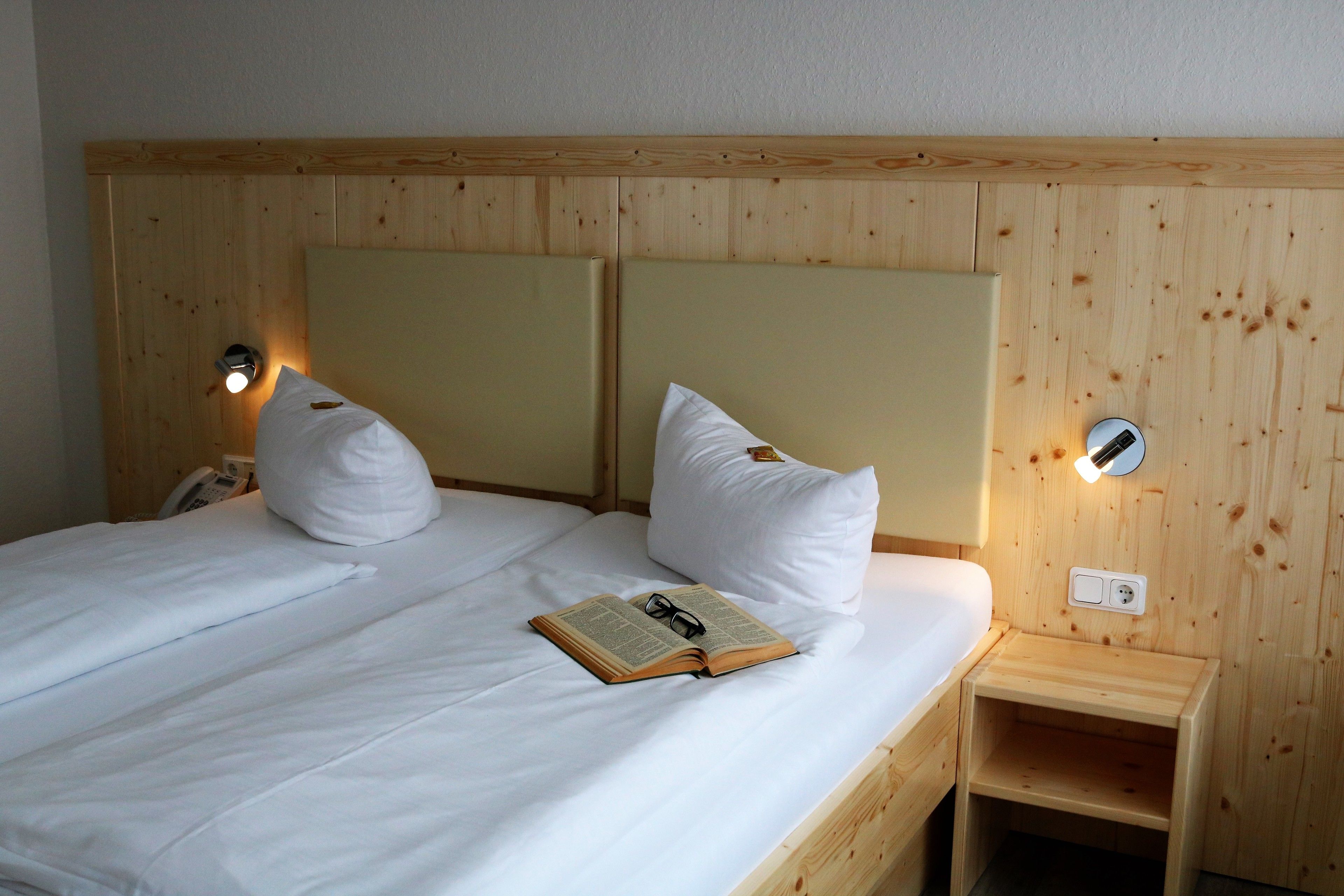 Un lit double avec du linge de lit blanc, deux oreillers, une table de chevet avec une lampe et un livre posé sur le lit.