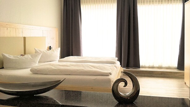 Una habitación de hotel con una cama, cabecera de madera, mesita de noche con un cuenco y vistas al exterior a través de amplias ventanas con cortinas.