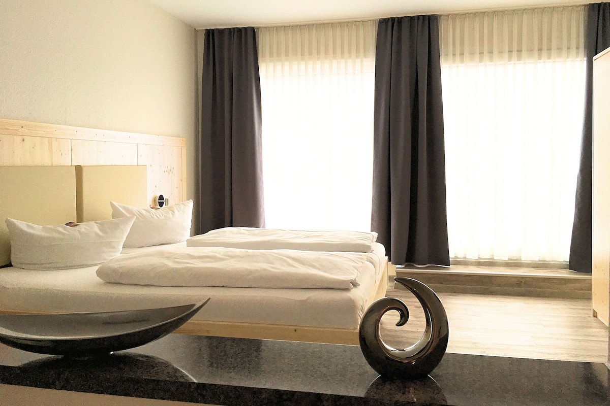 Ein Hotelzimmer mit einem Bett, einem hölzernen Kopfteil, einem Nachttisch mit einer Schale und Blick durch große, mit Vorhängen versehene Fenster nach draußen.