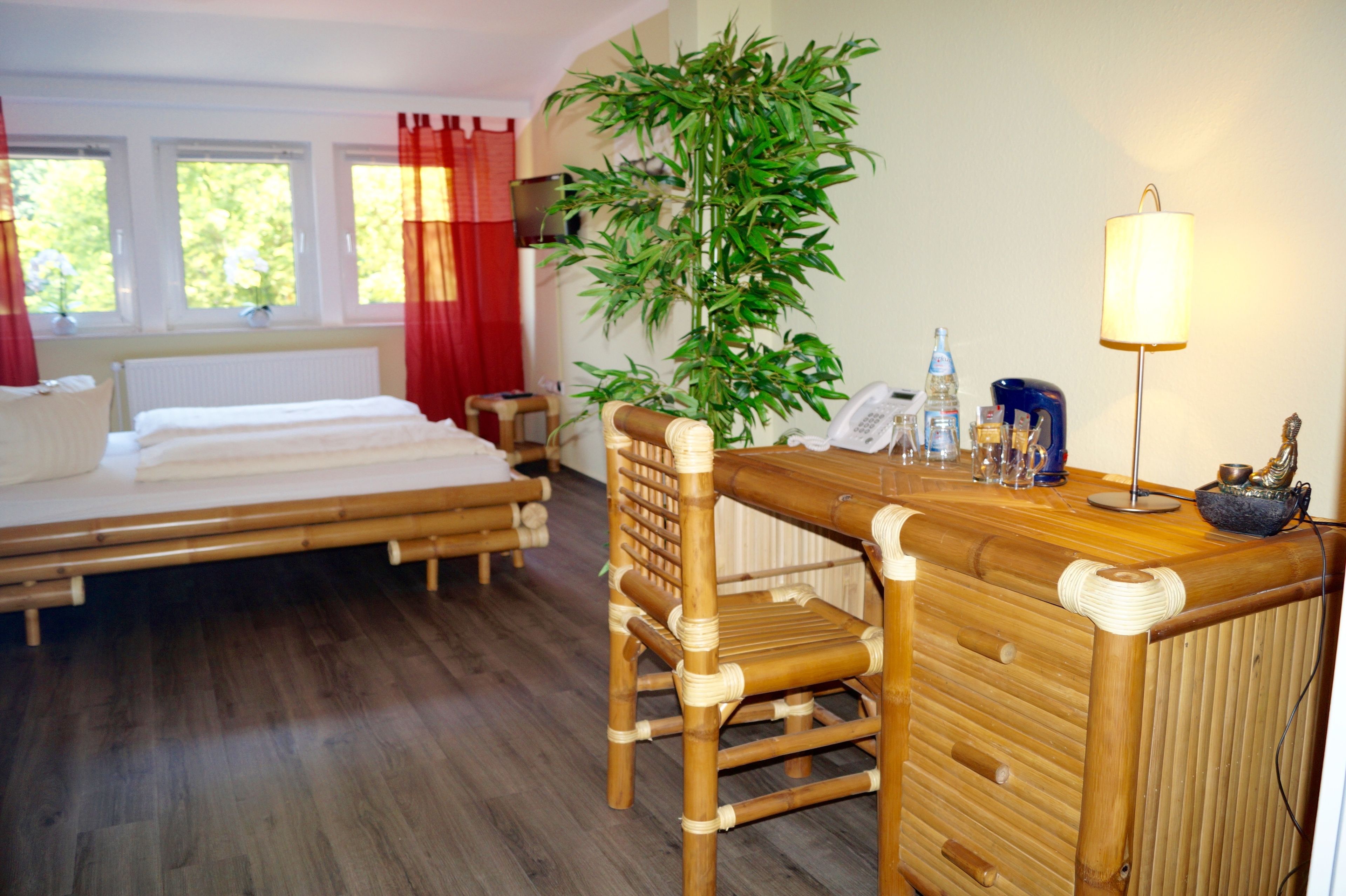 A room with a bed, a desk with a chair, a large plant, a lamp, and a telephone.