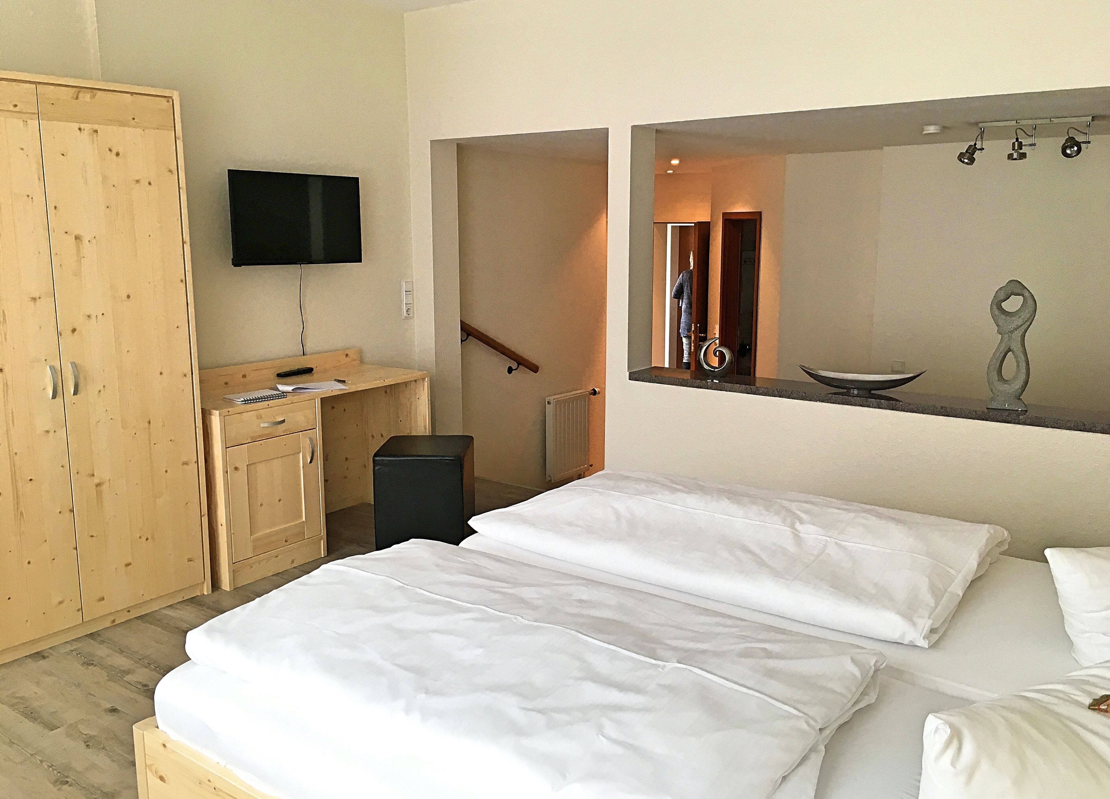 Habitación de hotel con un armario de madera, un escritorio y una silla, cama con ropa de cama blanca, televisor de pantalla plana montado en la pared y un estante con objetos decorativos.
