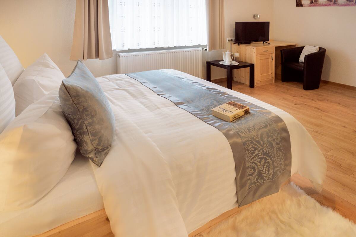 Ein ordentlich bezogenes Bett mit weißer und grauer Bettdecke, ein Buch auf dem Bett und ein Nachttisch mit Fernseher und Stuhl.