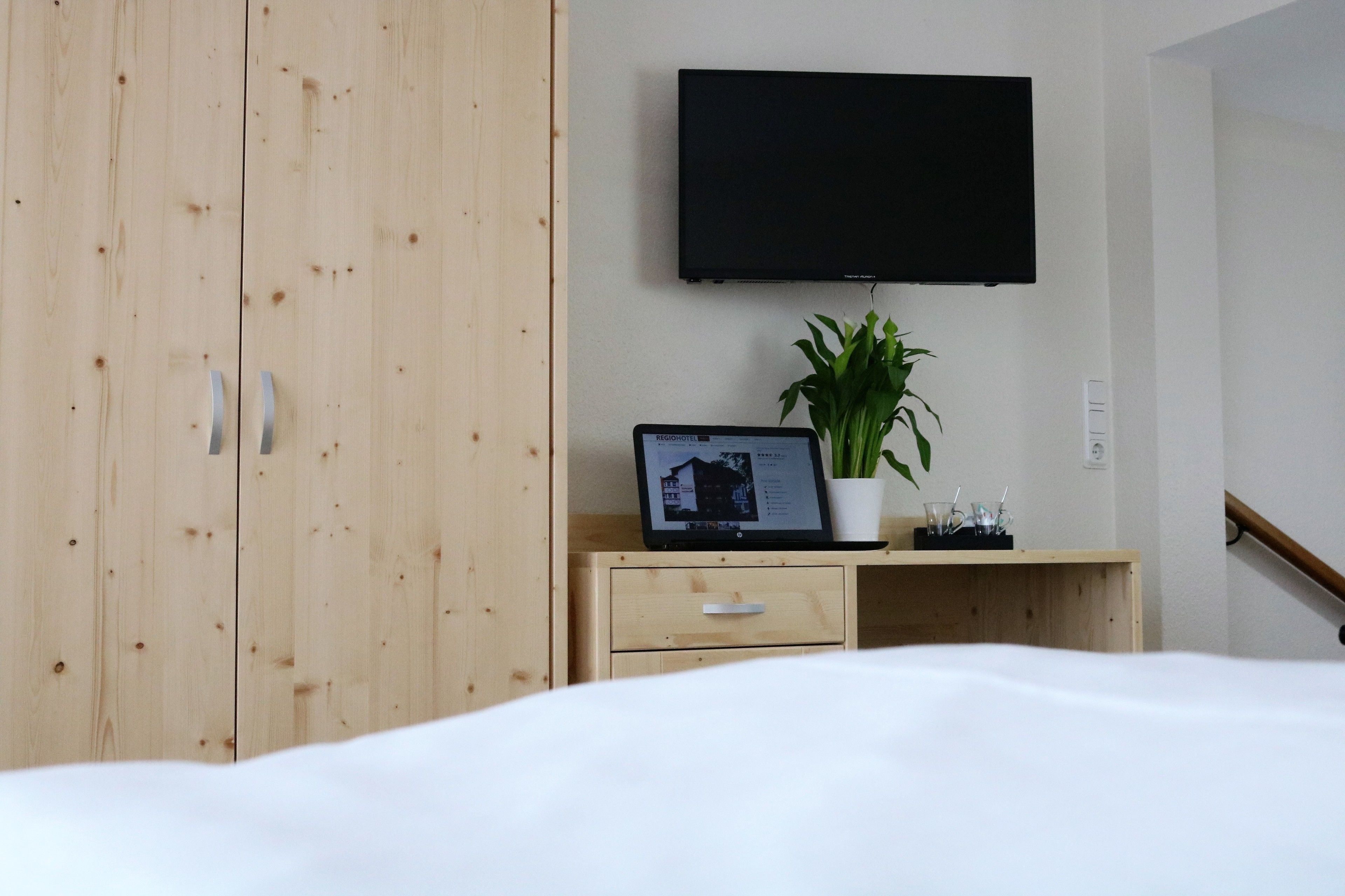 Een slaapkamer met een bed, een houten garderobe, een bureau met een laptop, een potplant en een televisie aan de muur.