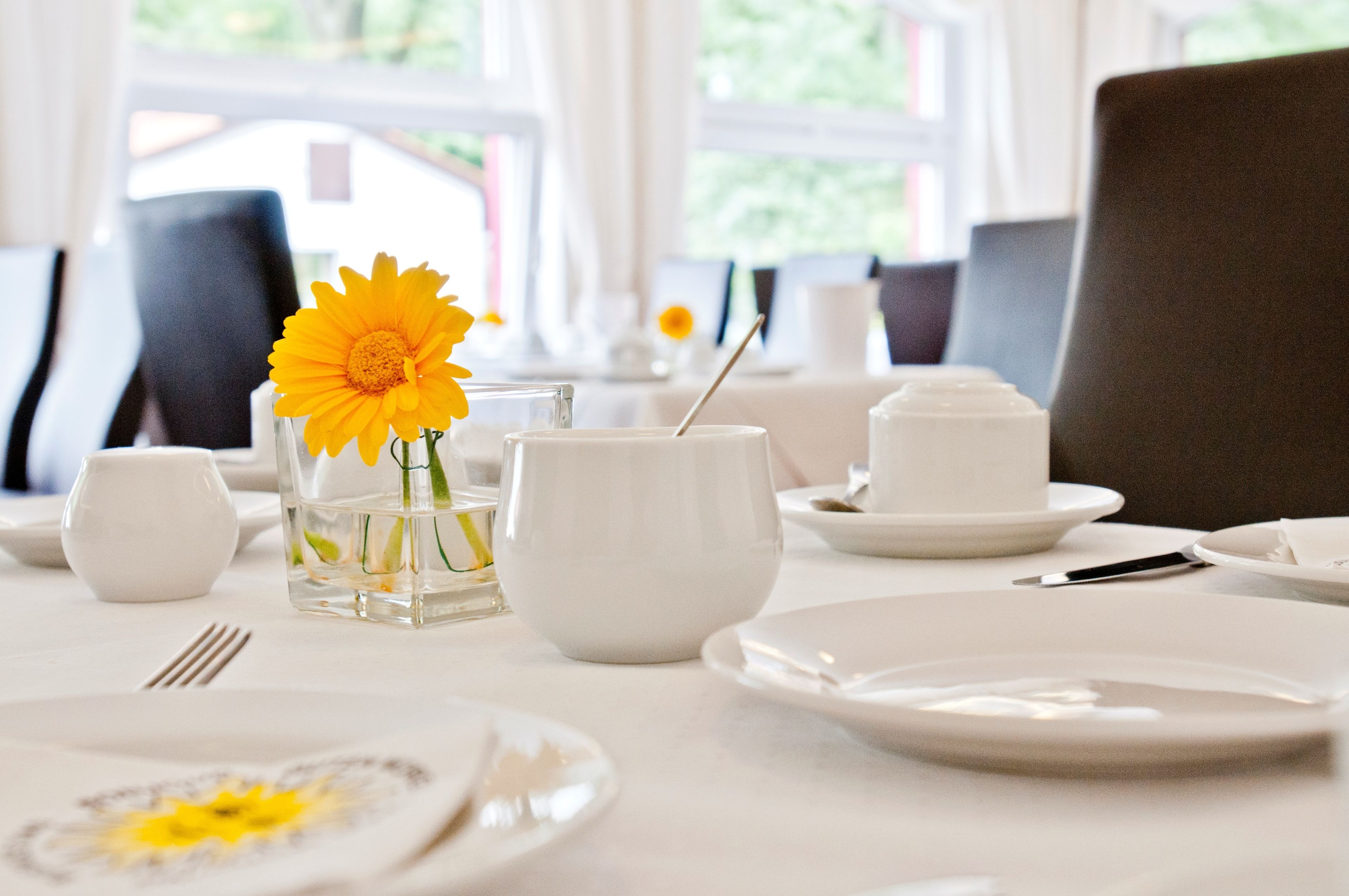 Una mesa de comedor dispuesta con platos blancos, tazones y un florero de vidrio con una flor amarilla.