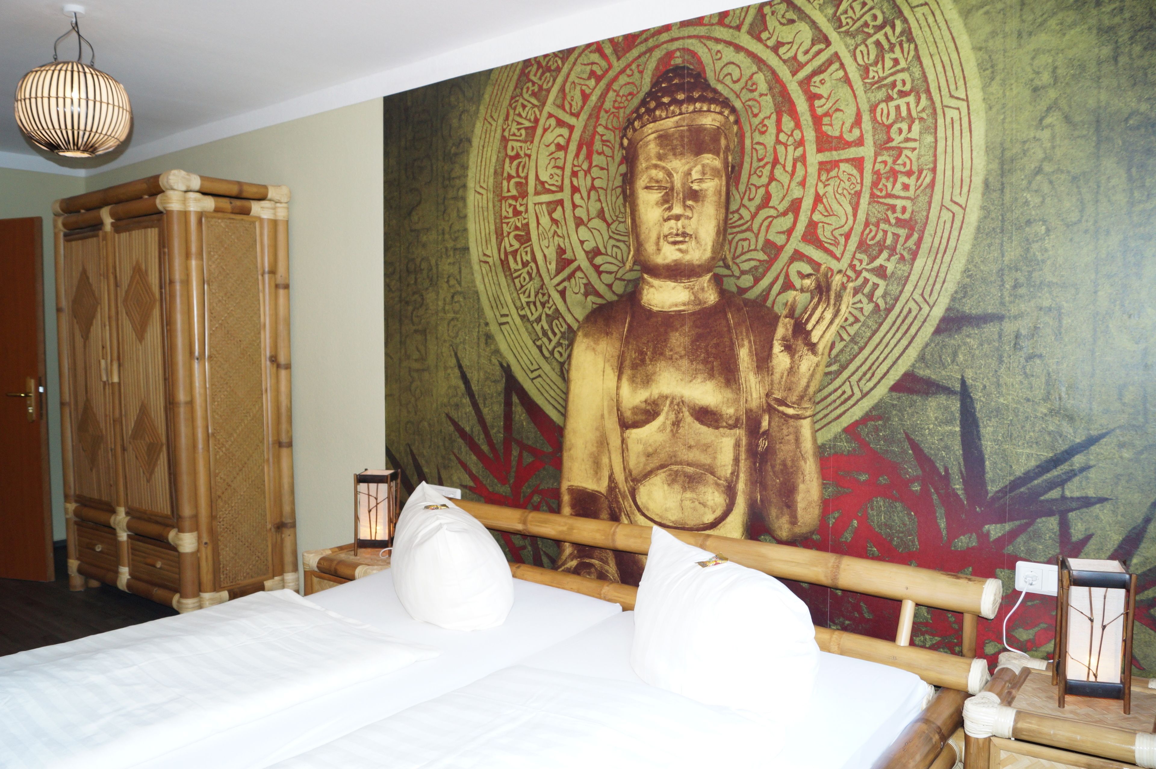 A room with a bed, a large Buddha mural, a wooden wardrobe, and a hanging light fixture.