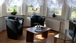 Una sala de estar moderna con un sofá de cuero negro, una mesa de centro de vidrio y una televisión. Amplias ventanas con cortinas translúcidas permiten la entrada de luz natural.