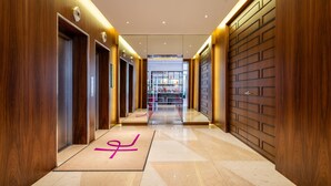 Un pasillo moderno de hotel con paneles de madera, un ascensor central y una vista despejada hacia la zona de recepción.