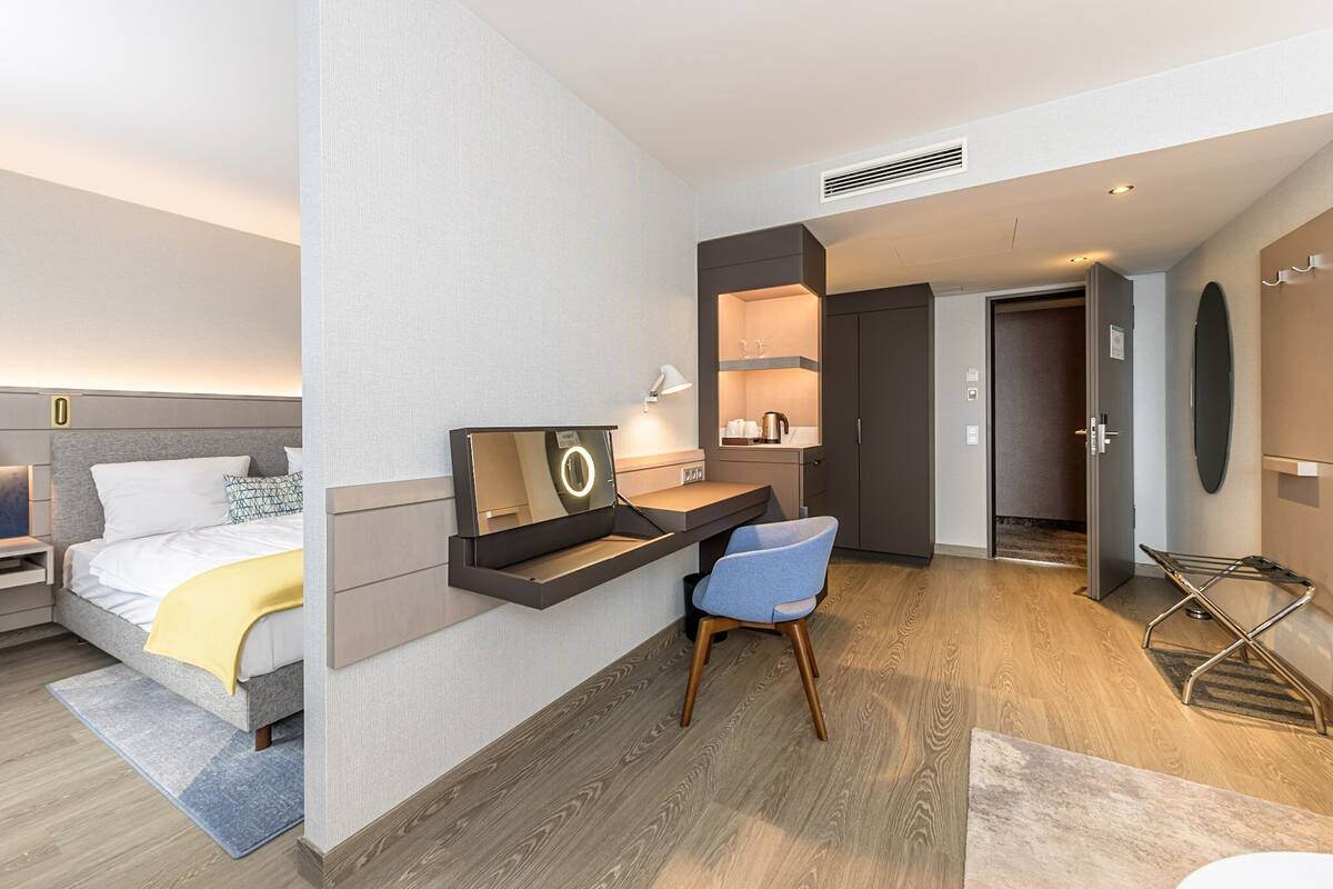 A modern hotel room with a bed, desk, chair, and built-in storage.