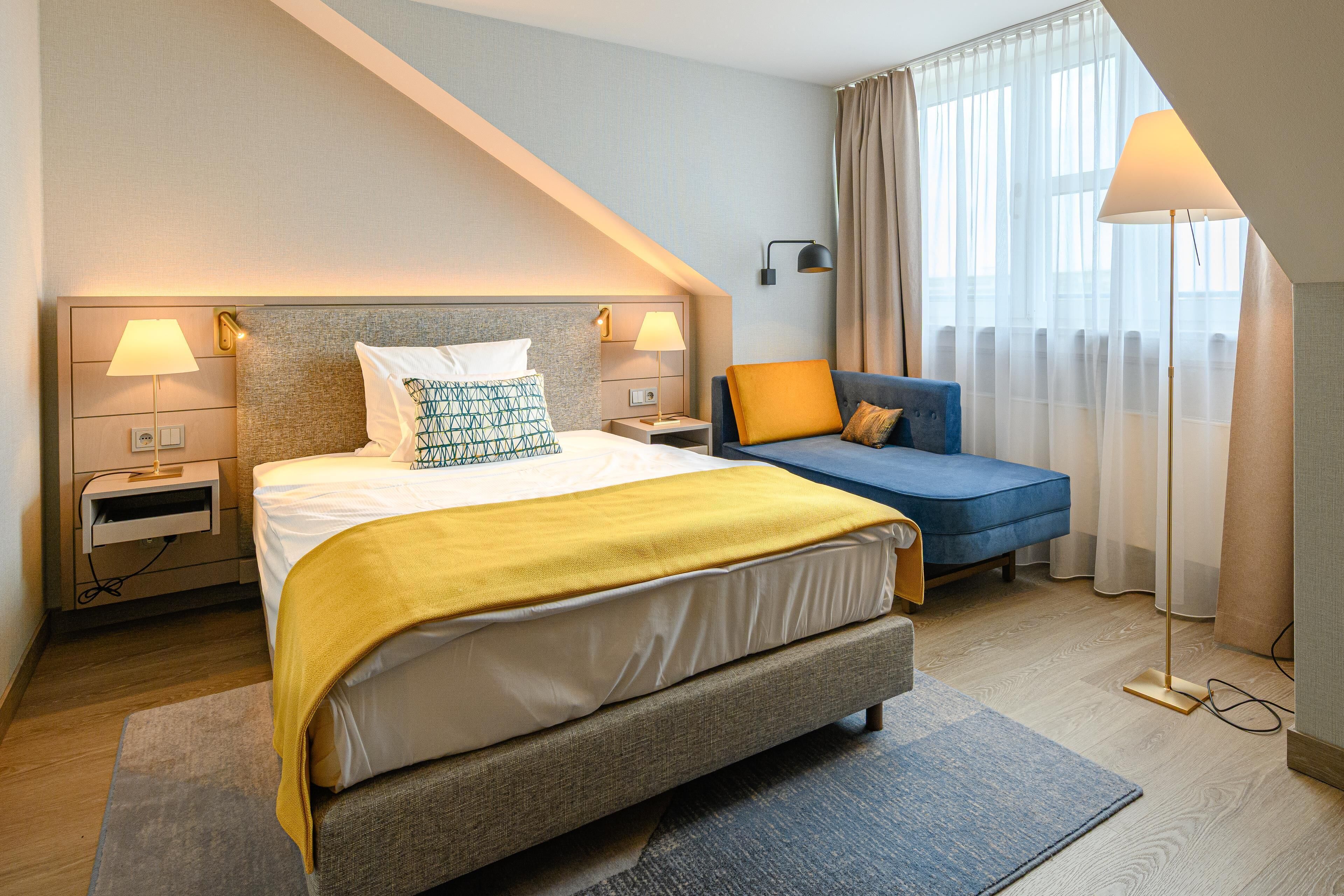 A hotel room with a bed, bedside tables, a sofa, a lamp, and a view of the outside.