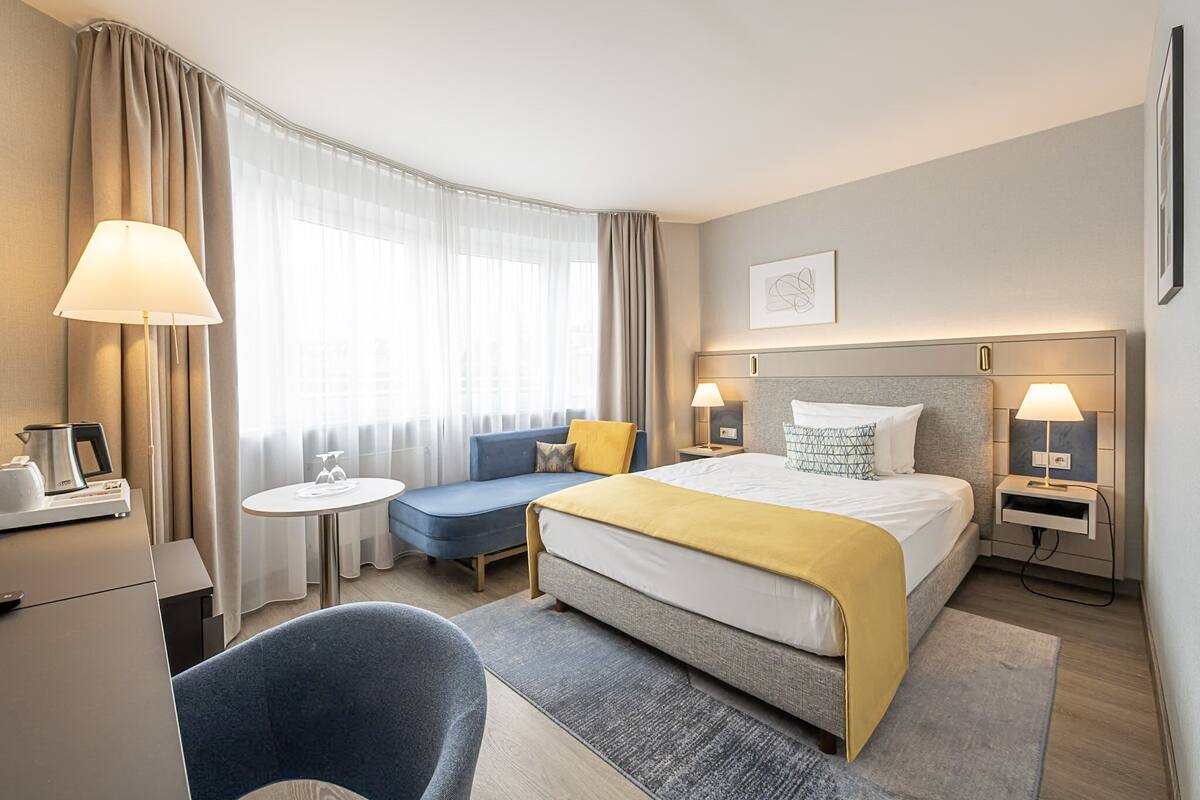 A hotel room with a bed, bedside tables, a desk, a chair, a sofa, and a small table.