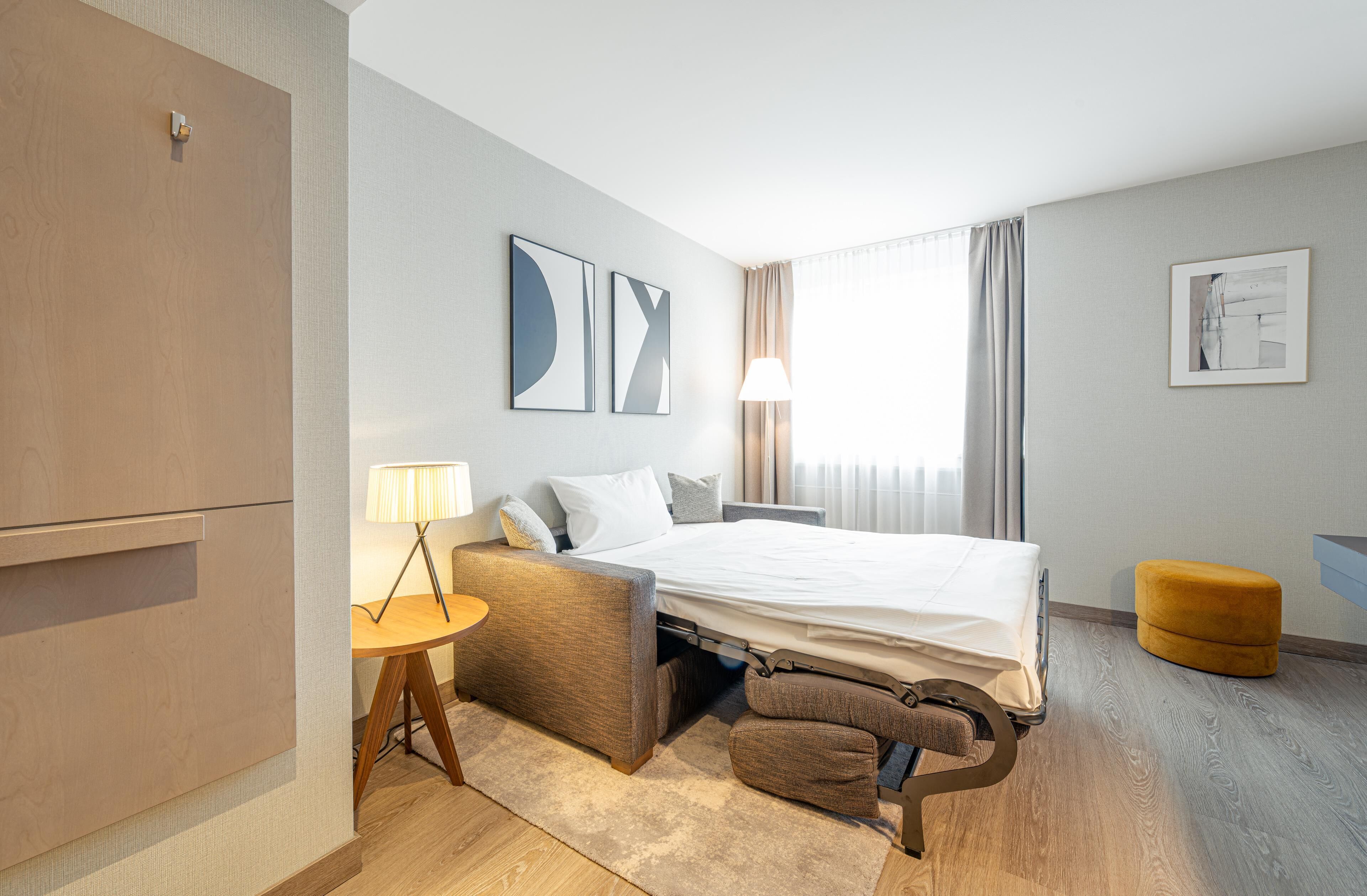 Une chambre d’hôtel moderne équipée d’un lit, d’un bureau, d’une chaise, d’une lampe et d’un petit pouf.