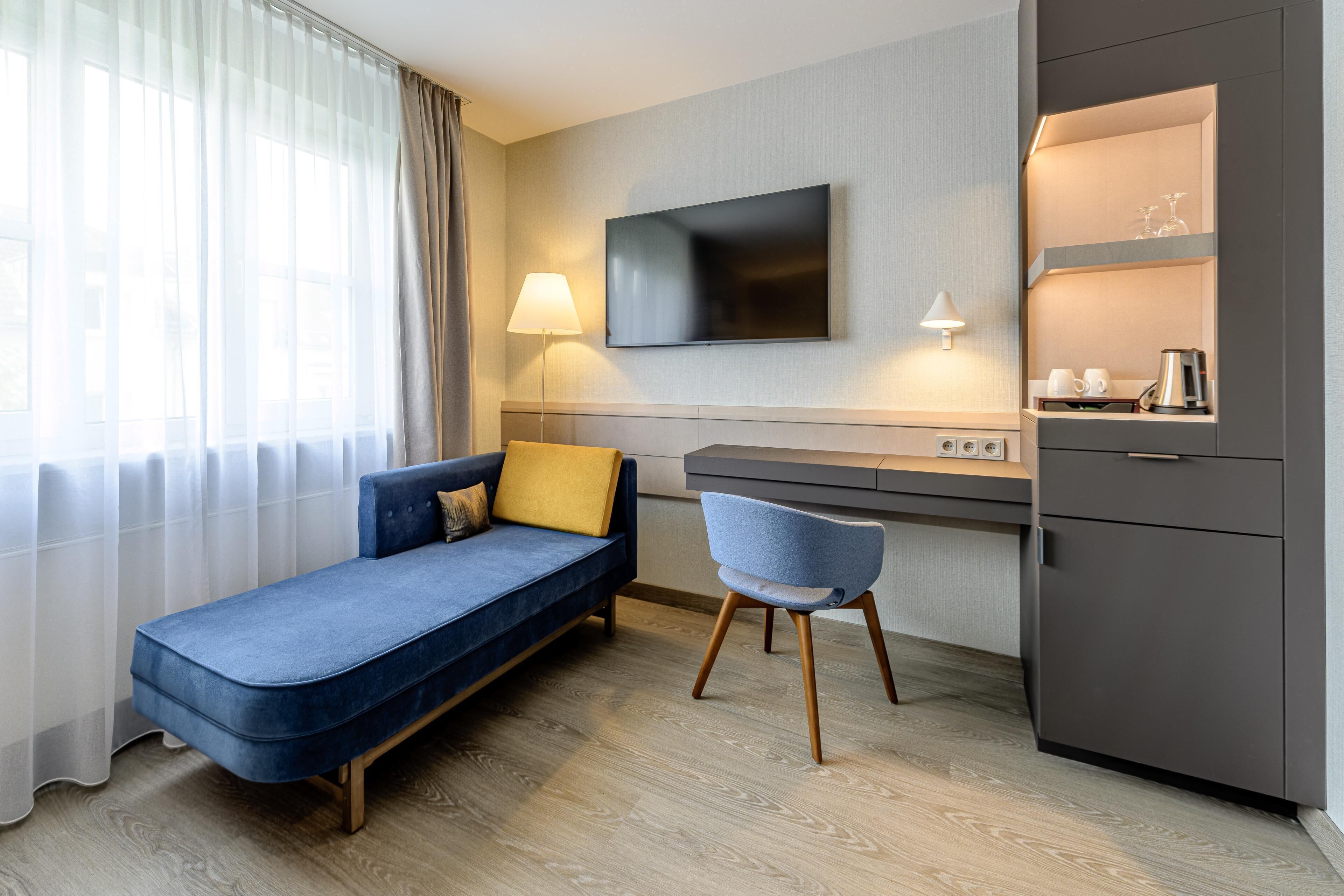A modern hotel room with a sofa, a desk, a chair, a TV, a lamp, and a minibar.