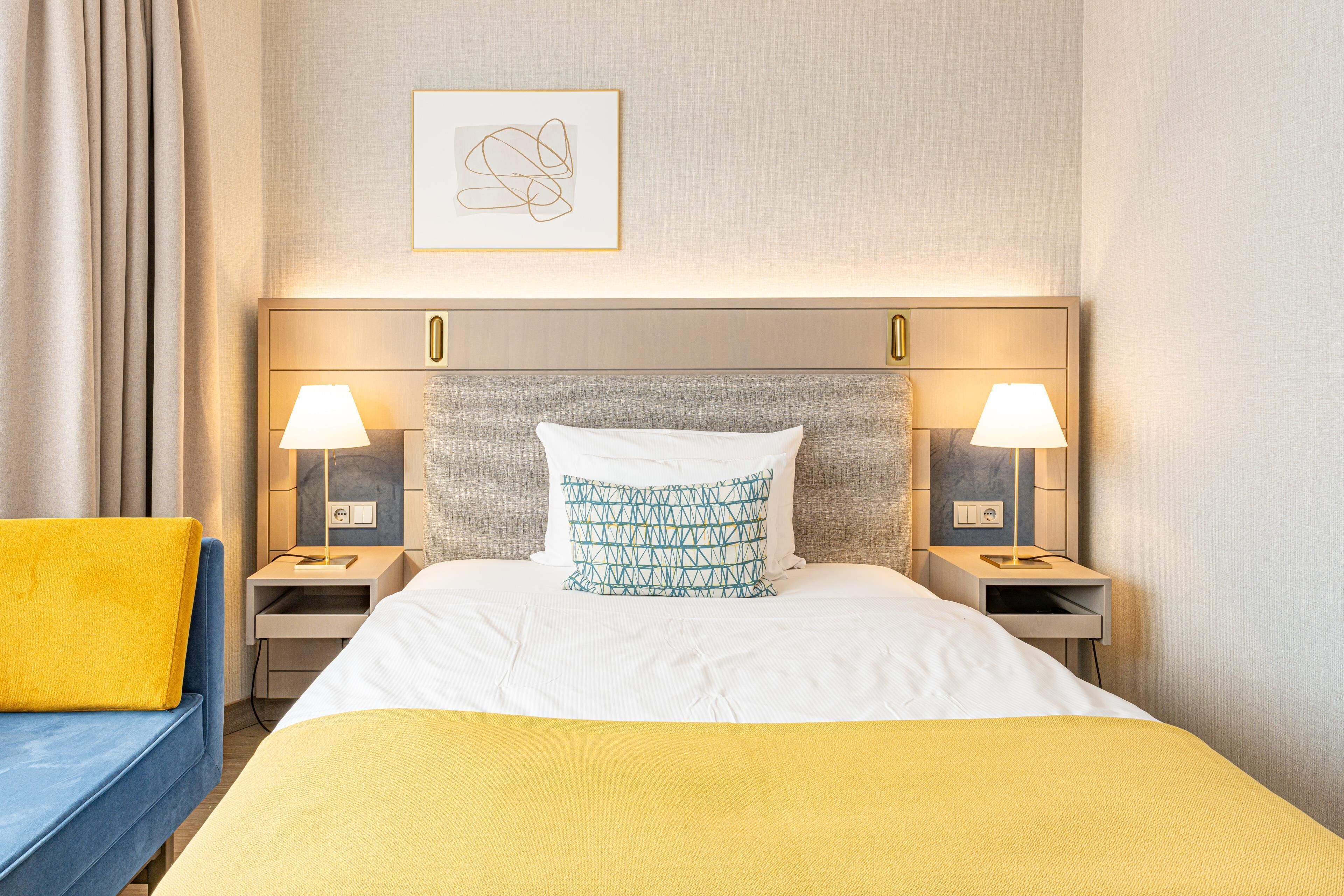 Habitación de hotel con cama, dos mesitas de noche con lámparas, un cuadro en la pared y una manta amarilla.
