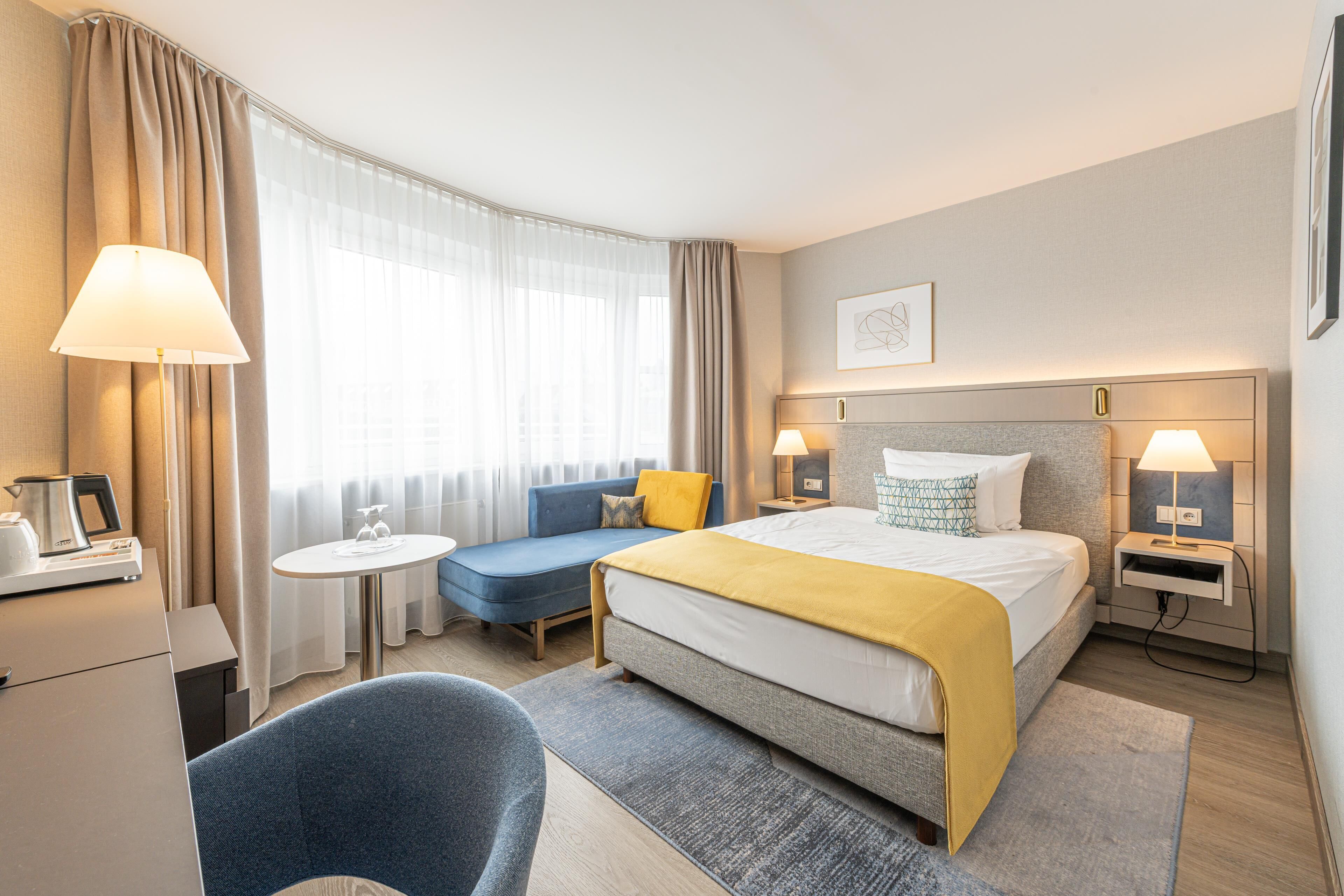 Une chambre d’hôtel comprenant un lit, des tables de chevet, un bureau, une chaise, un canapé et une fenêtre avec des rideaux.