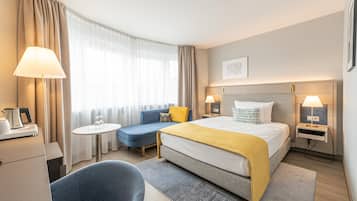 A hotel room with a bed, bedside tables, a desk, a chair, a sofa, and a window with curtains.