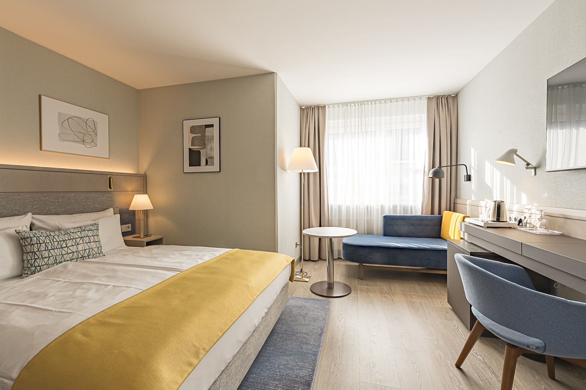 A hotel room with a bed, a desk, a chair, a round table, and a large window with curtains.