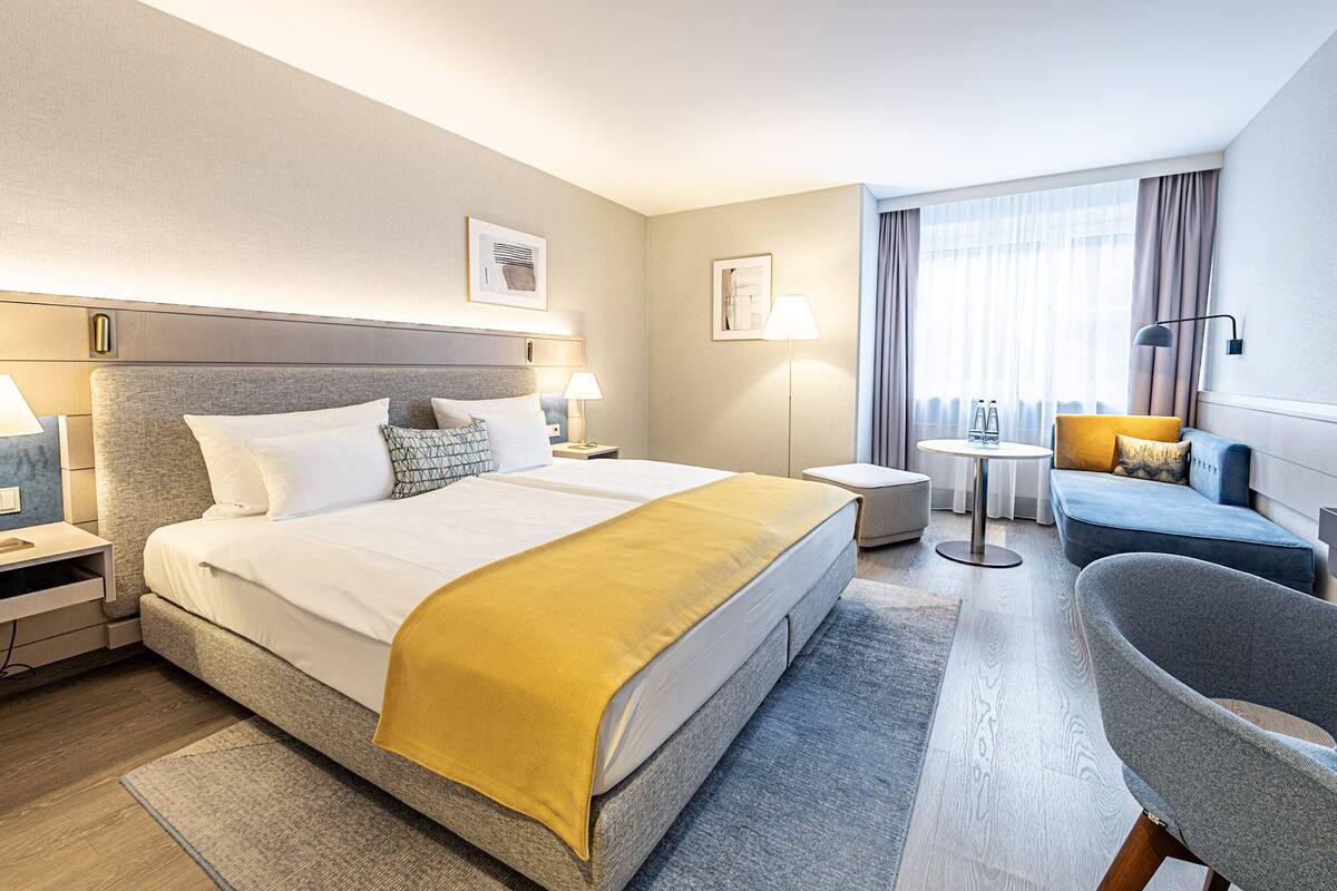 A hotel room with a large bed, a small table, a sofa, and a chair.