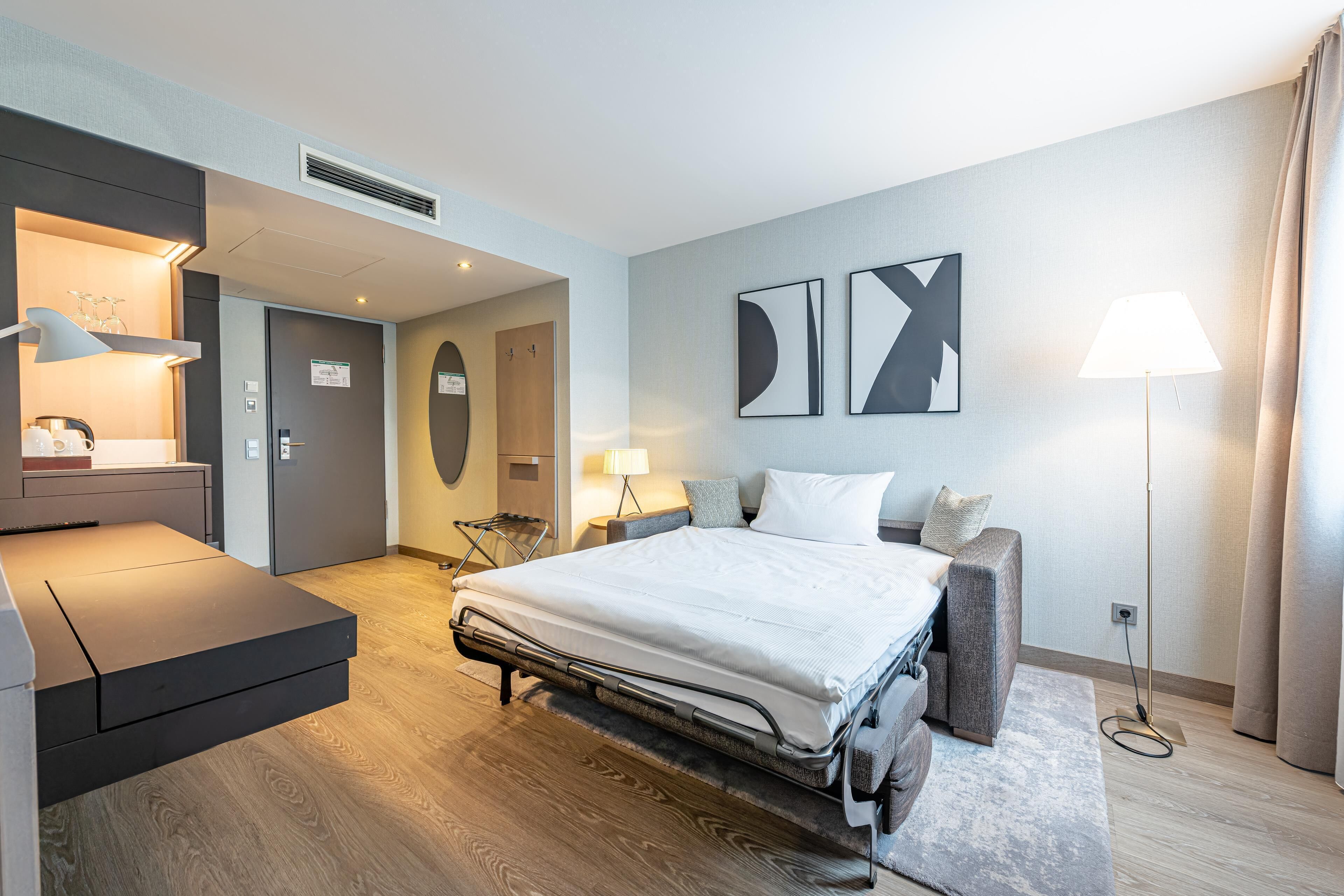 A modern hotel room with a bed, a desk, a chair, a sofa, and a lamp.