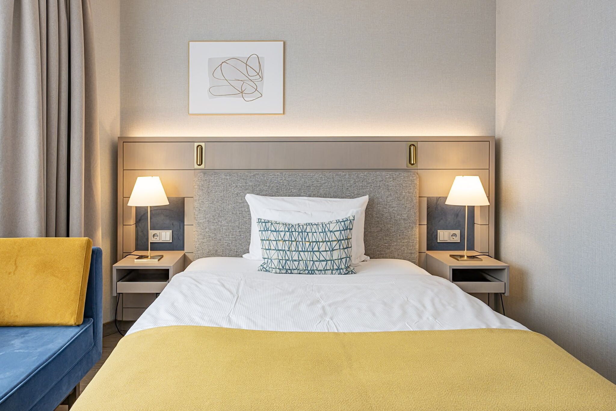 A hotel room with a bed, two bedside tables with lamps, a framed artwork on the wall, and a yellow throw blanket.