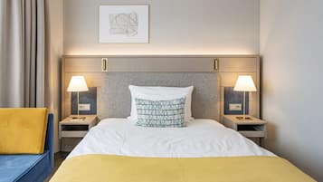 A hotel room with a bed, two bedside tables with lamps, a framed artwork on the wall, and a yellow throw blanket.