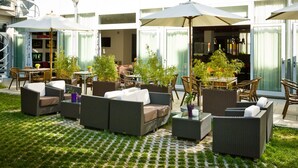 Un espacio al aire libre con mobiliario de mimbre y sombrillas, rodeado de plantas en macetas y un piso de baldosas.