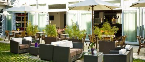 An outdoor seating area with wicker furniture and umbrellas, surrounded by potted plants and a tiled floor.