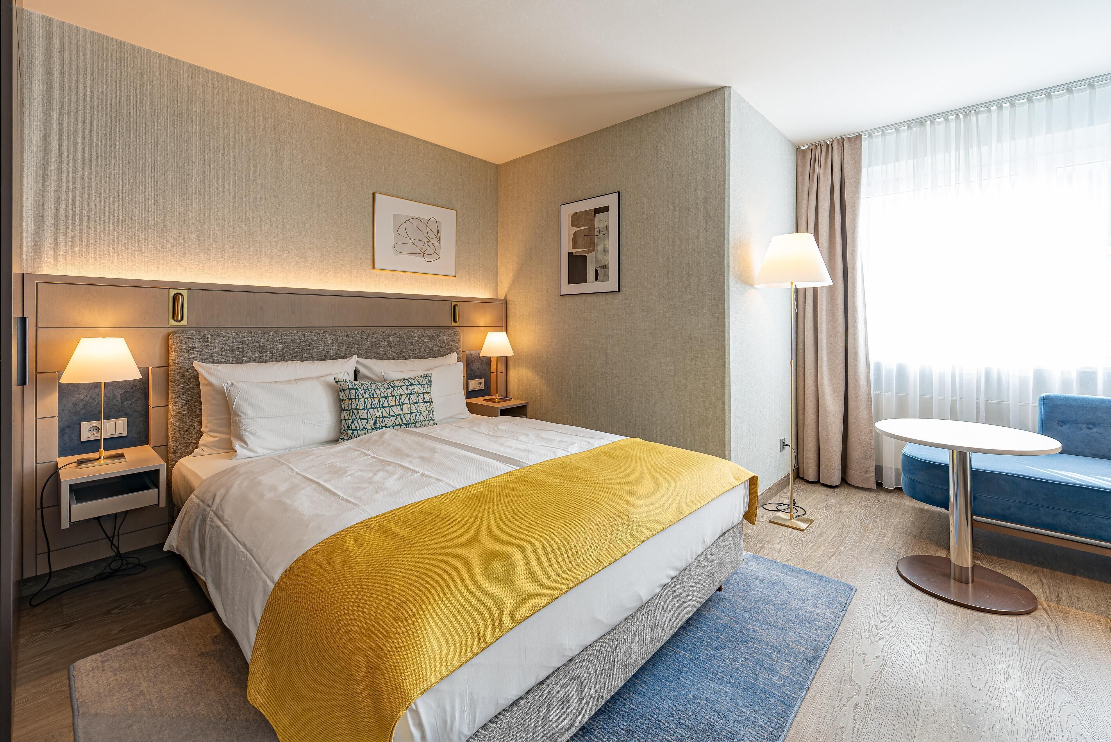 A hotel room with a bed, bedside tables with lamps, a round table, a sofa, and a view of the city.