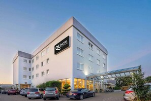 Un hôtel moderne, avec une enseigne bien visible, plusieurs étages et un parking où sont stationnées plusieurs voitures.