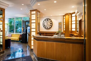 A hotel reception area with a large vintage clock, a reception desk, and a view of greenery outside.
