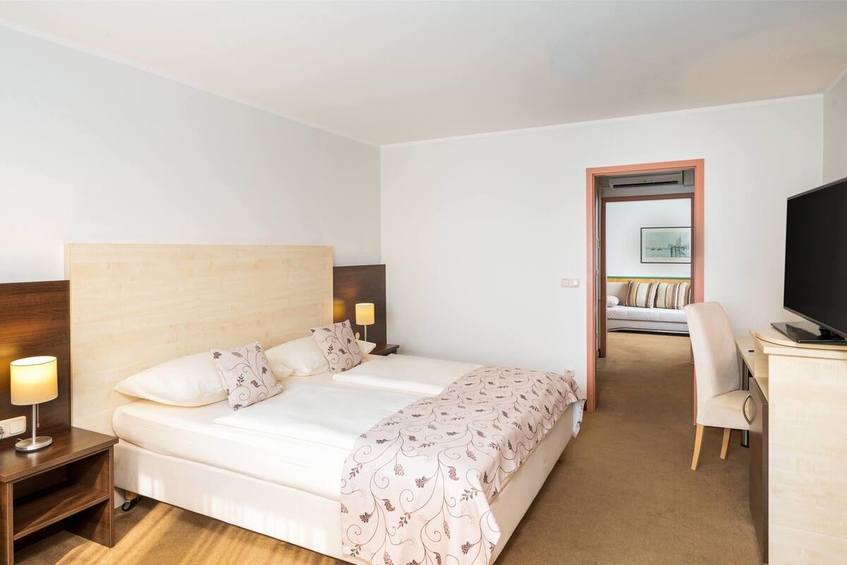 Habitación de hotel con una cama grande, lámparas de noche, un escritorio con televisión de pantalla plana y vista a otra habitación por una puerta abierta.