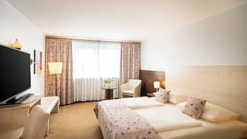 Ein Hotelzimmer mit einem Bett, einem Schreibtisch mit Fernseher, einem Sessel, einem kleinen Tisch mit Lampe und einem Fenster mit Vorhängen.
