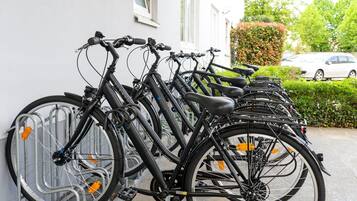 En rad cyklar står parkerade längs en vägg, med en bil och grönska i bakgrunden.