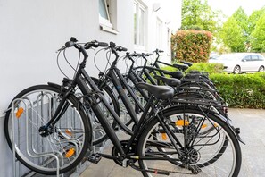 Une rangée de vélos est garée contre un mur, avec une voiture et de la verdure en arrière-plan.