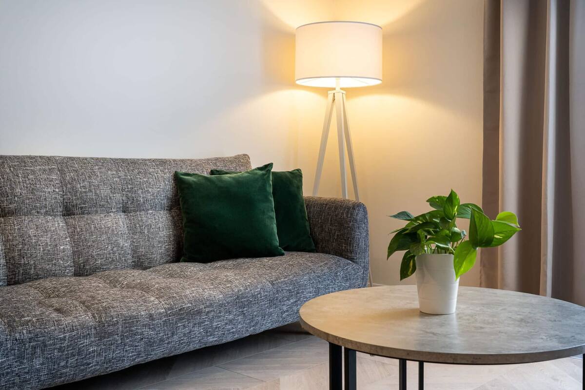 Un sofá gris con un cojín verde, una lámpara de pie blanca y una mesita con una planta en maceta.