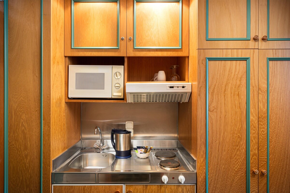 Een kleine keuken met houten kastjes, een magnetron, een waterkoker en een fornuis.
